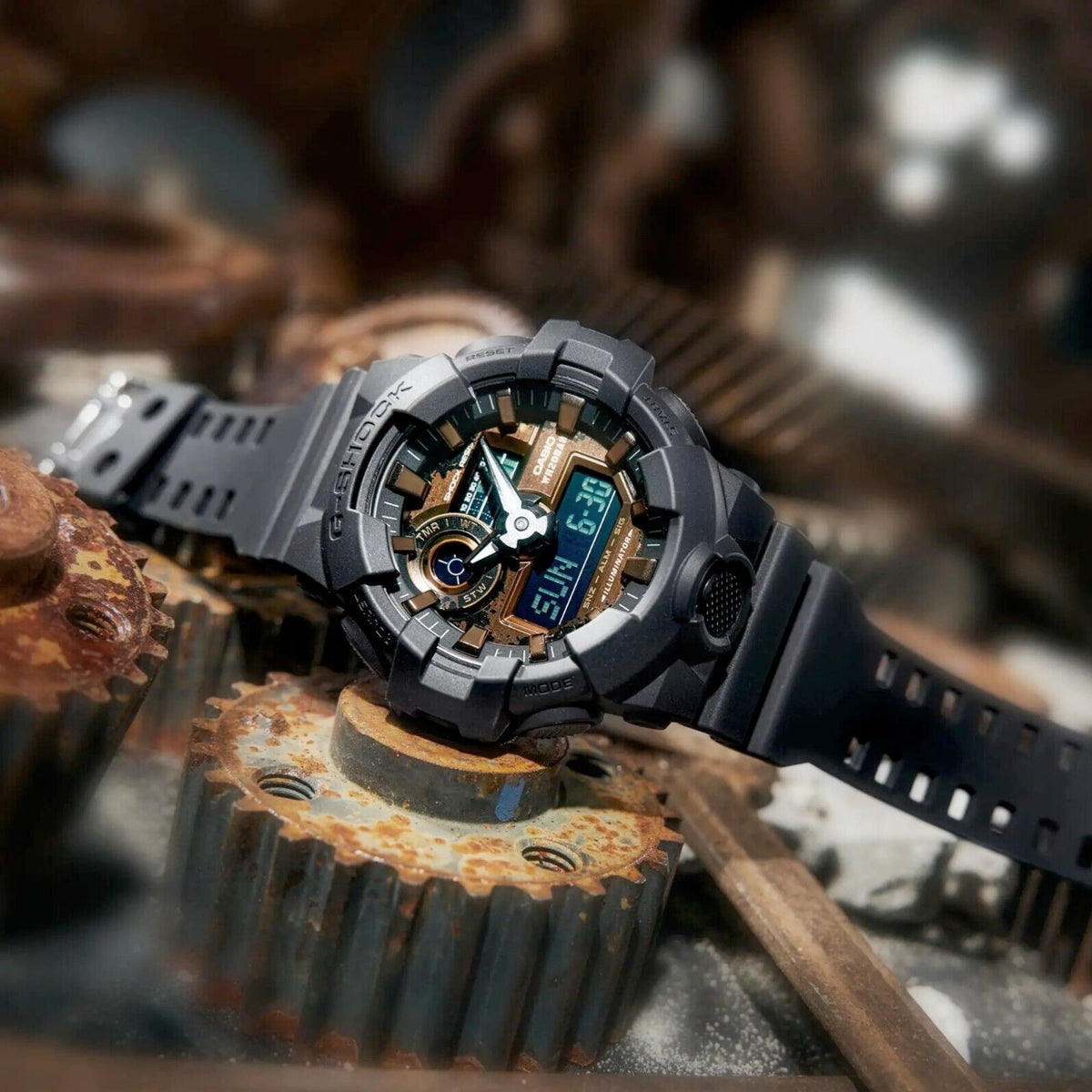 Men's black G-SHOCK digital watch with gold bezel ring, resin strap; angled side view on metal.