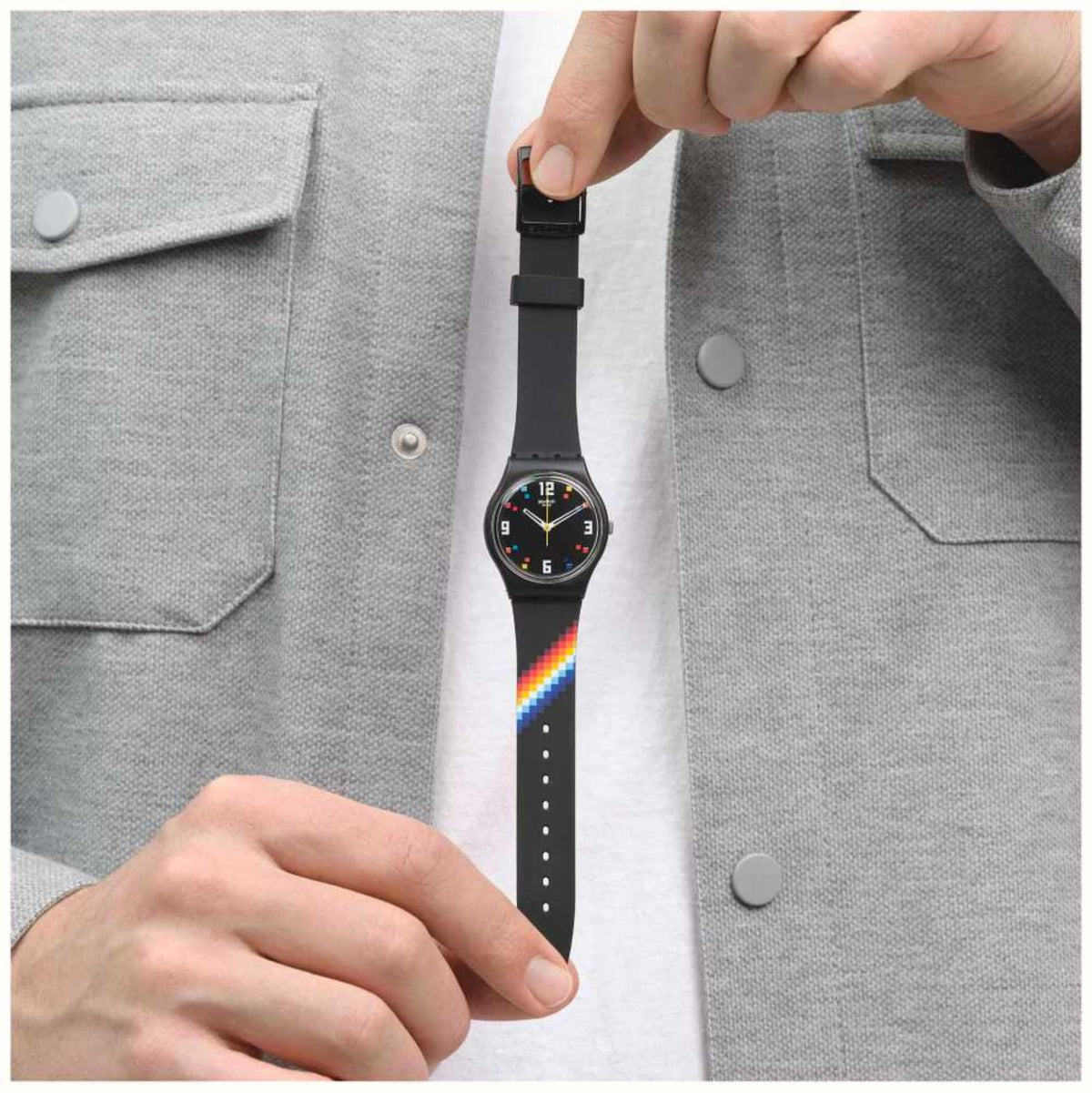 Hand holding black Swatch wristwatch; black strap with rainbow stripe and colorful dial.