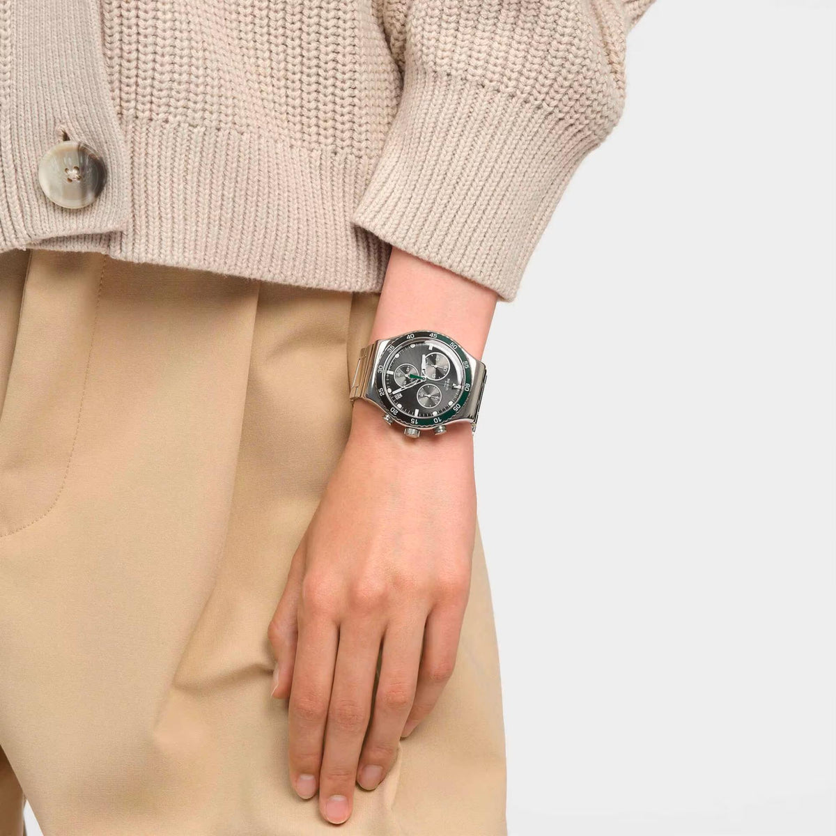 Stainless steel wristwatch on wrist with dark face and green bezel, over beige trousers