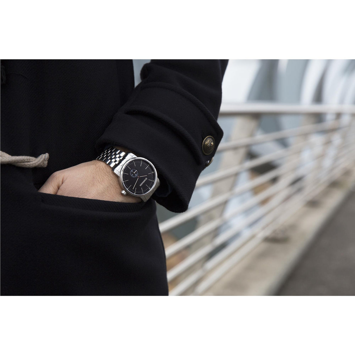 Watch on wrist in dark jacket: black dial, silver hands, and linked stainless bracelet.