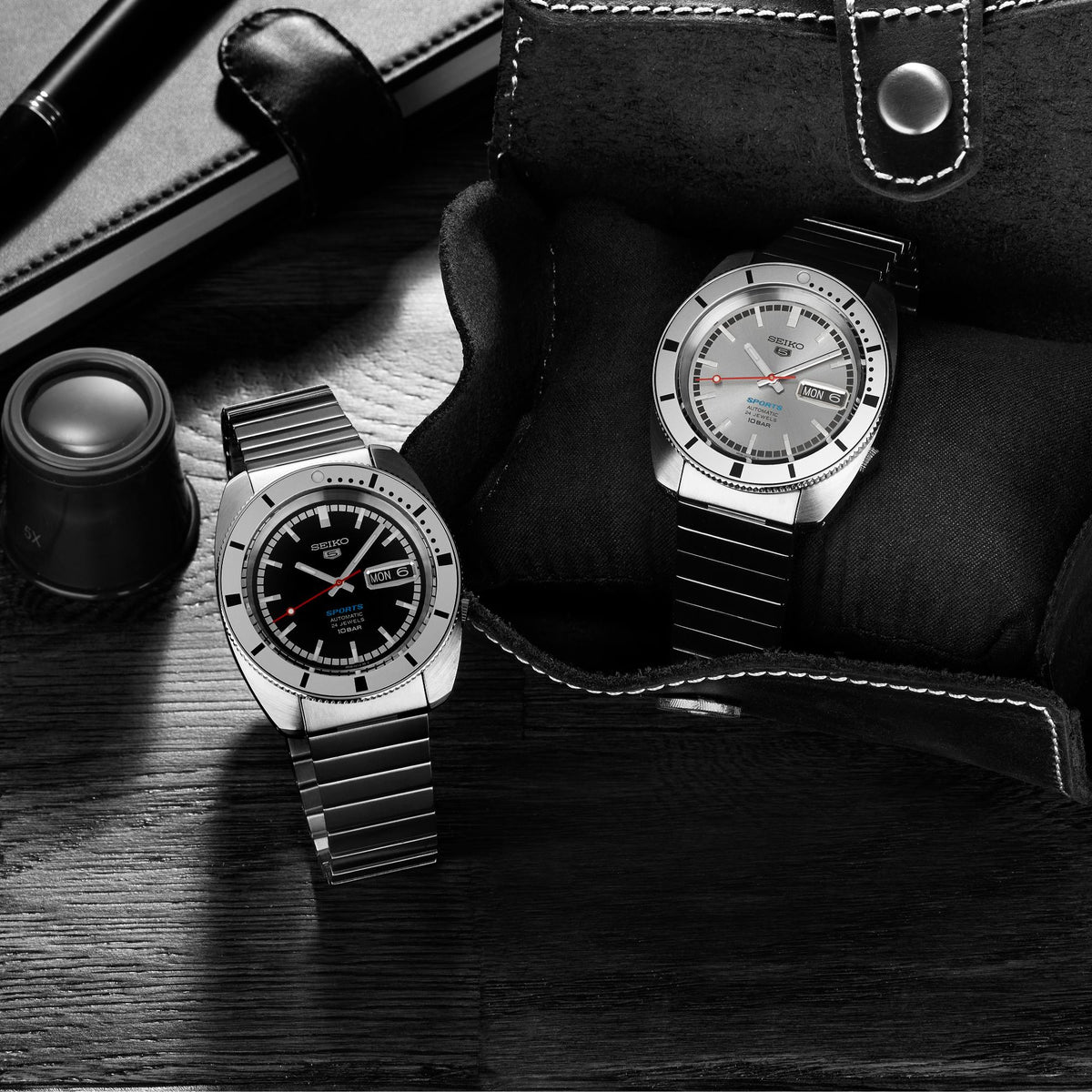 Two men's stainless steel watches on dark leather and wood; metal bracelets and light dials.
