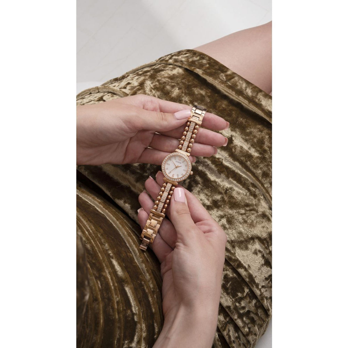 Gold-tone watch with white dial and beaded bracelet, shown in hands near a sleeve