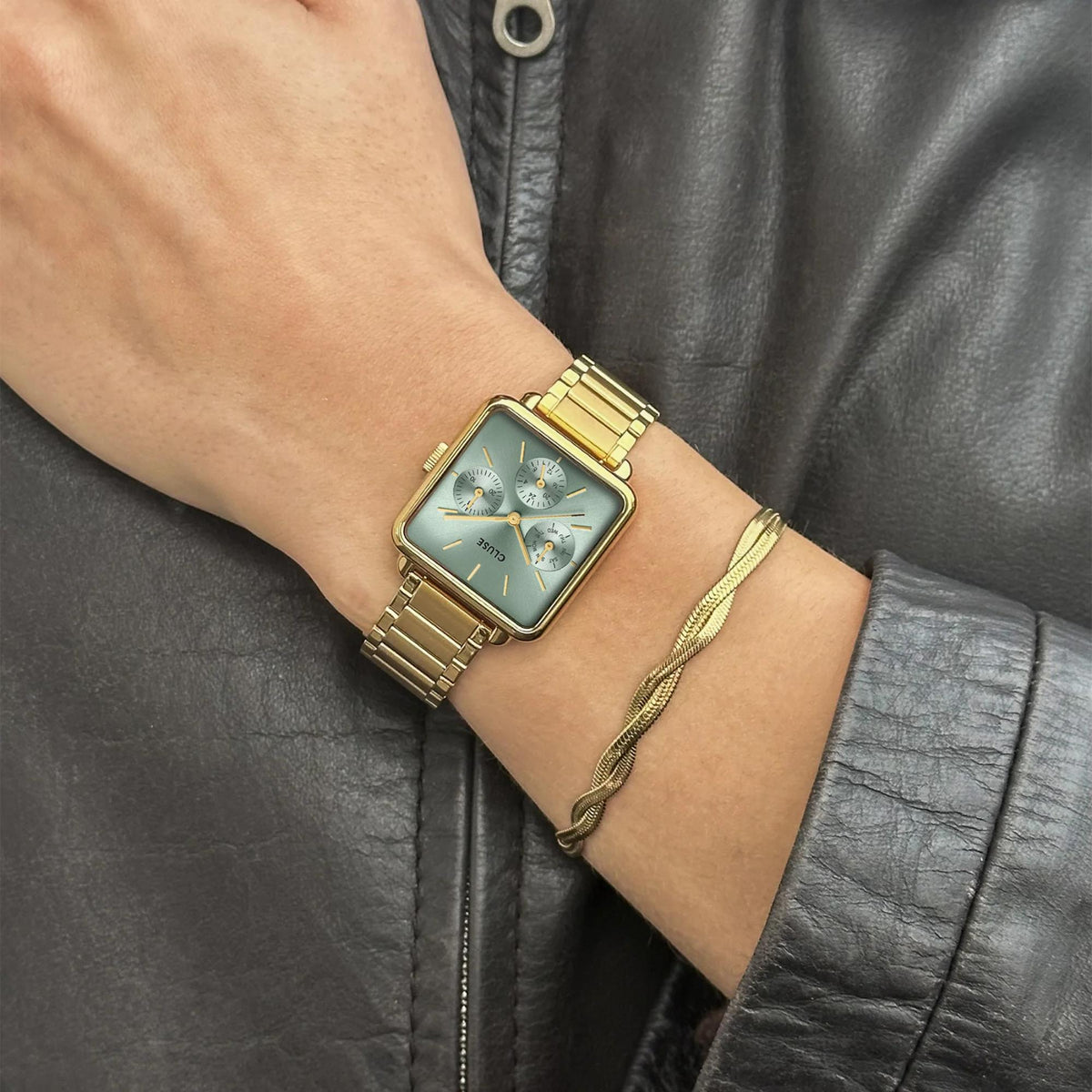 Gold-tone steel wristwatch with teal-green dial on wrist against leather jacket