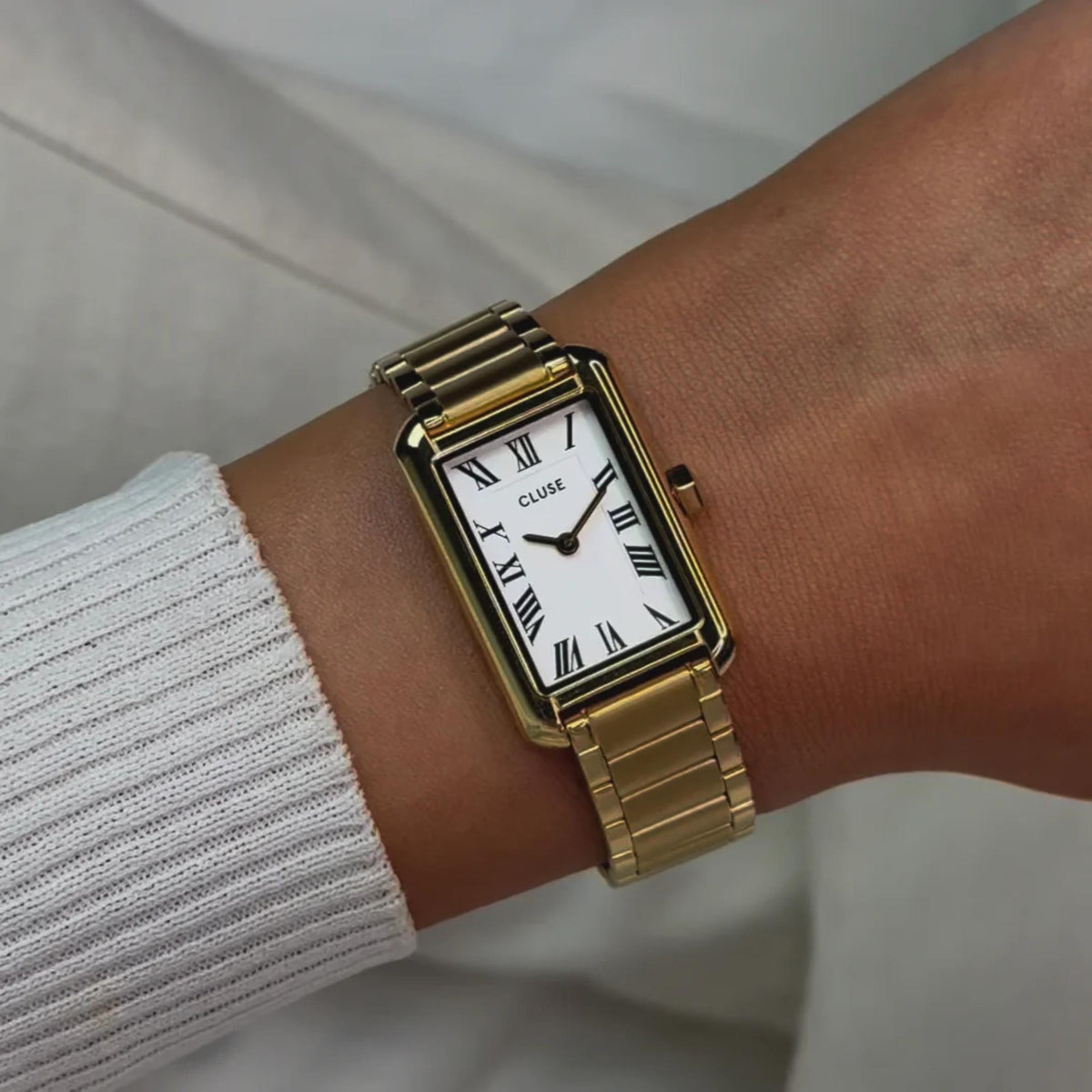 Gold-tone rectangular watch with white dial and roman numerals; metal link bracelet on wrist