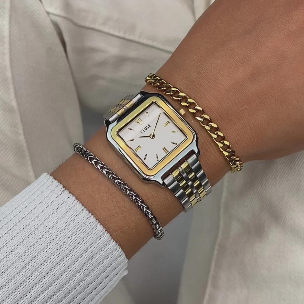 Two-tone square watch with white dial on wrist with braided charm bracelet