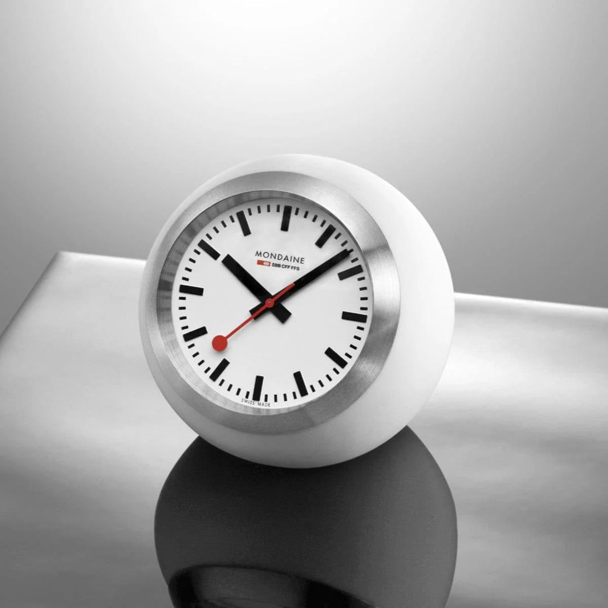 Mondaine Globe Table Clock A660.30335.16SBA resting on a reflective chrome surface with a crisp white dial and black markers