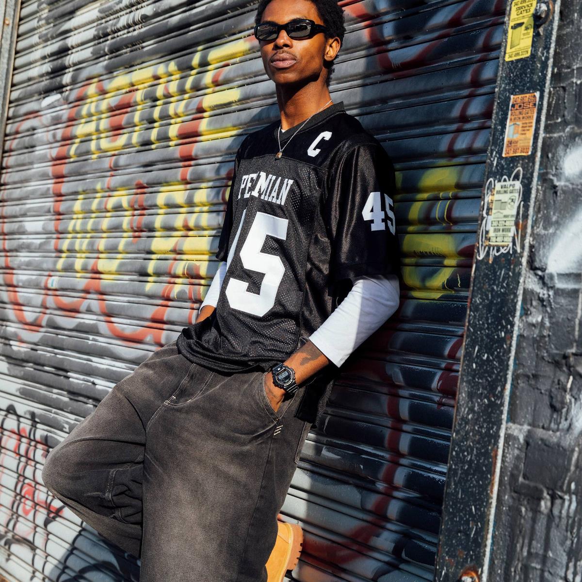 Model wearing the Casio G-Shock 6900 Series DW-6900RL-1ER black resin watch styled with athletic streetwear in front of a metal shutter