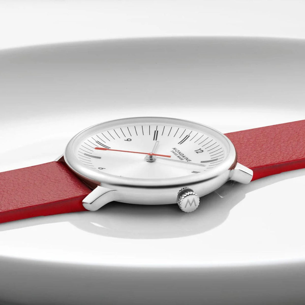 Side view of Mondaine watch resting flat, highlighting the ultra-slim polished silver watch case, the textured crown engraved with the brand's 'M' logo, and the red leather strap extending outward