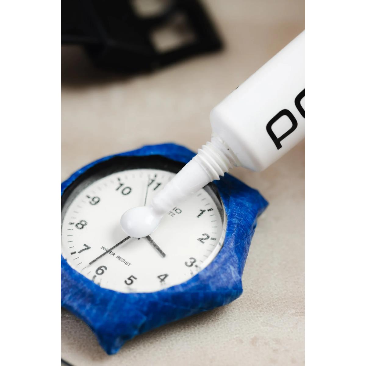 Scratch remover polish tube dispensing white polish onto watch on blue pad.