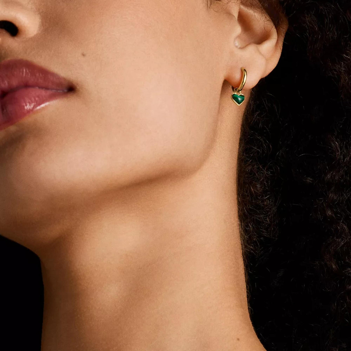 Gold hoop earrings with dark green heart charm, worn on model's ear