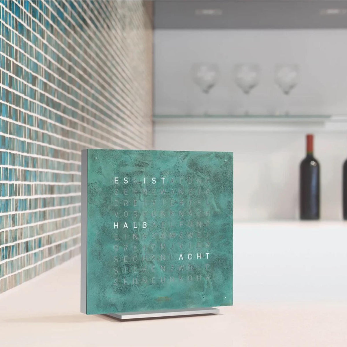 Turquoise square word clock with glowing letter display on a tabletop stand.