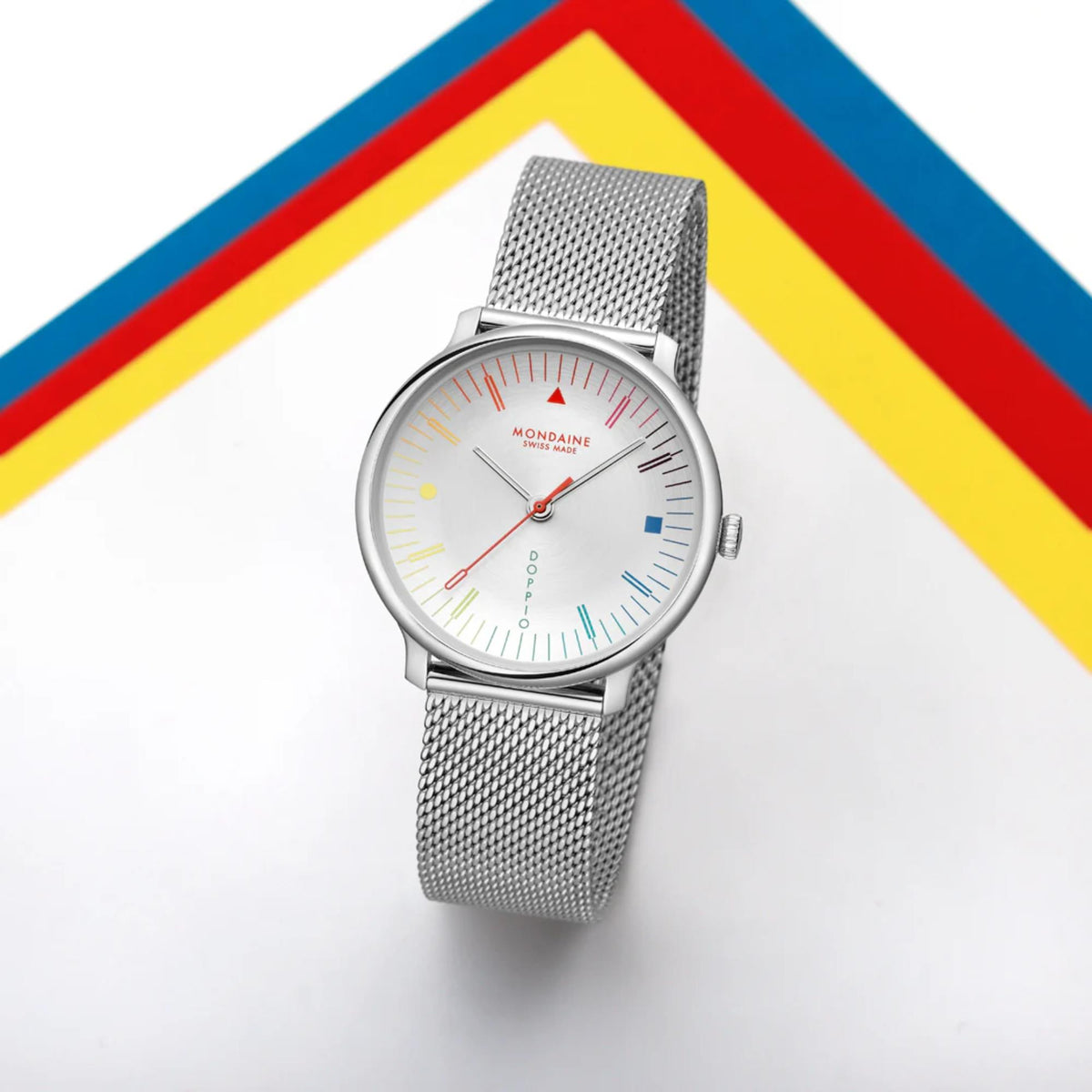 Mondaine Doppio quartz watch resting at an angle on a clean white surface with bold primary blue, red, and yellow stripes in the background