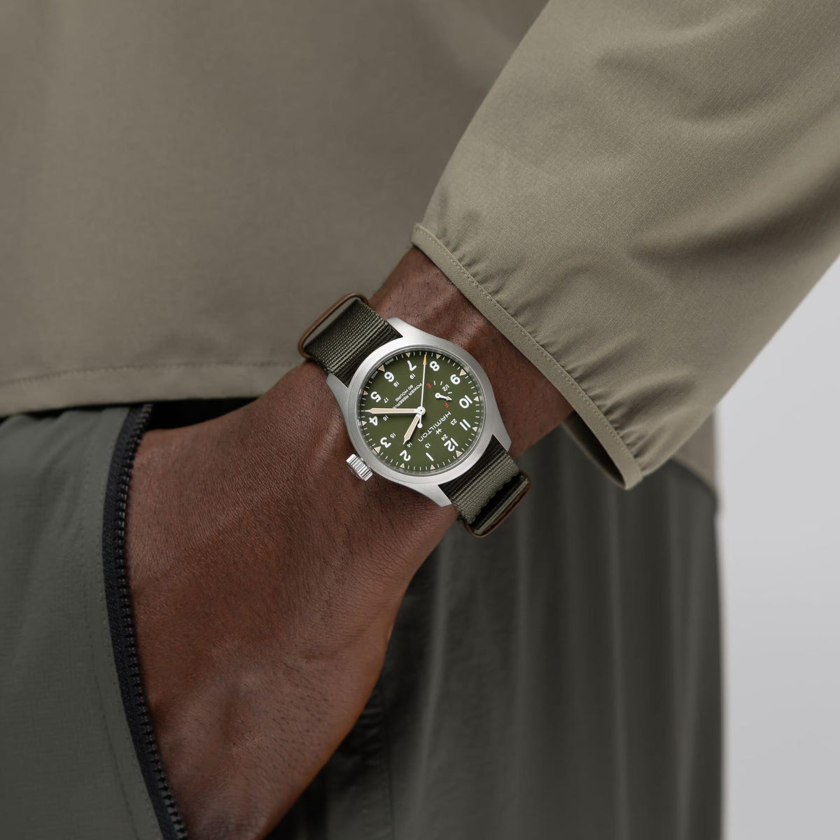 Men's field watch on wrist with green dial and olive NATO strap under khaki jacket
