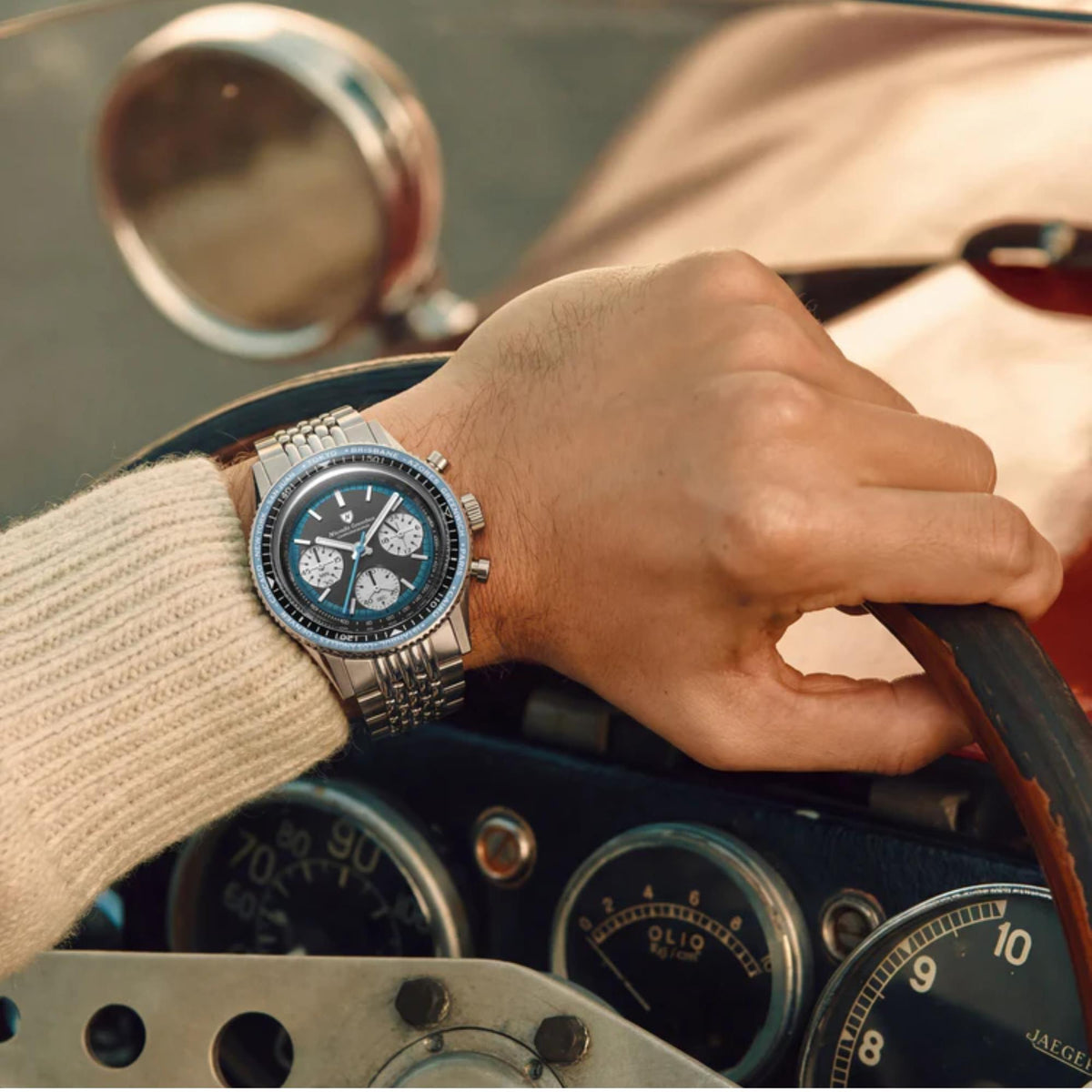 Close-up of Nivada Grenchen Chronoking watch 87053Q04 with a blue bezel worn on a person's wrist, who is holding the steering wheel of a vintage car