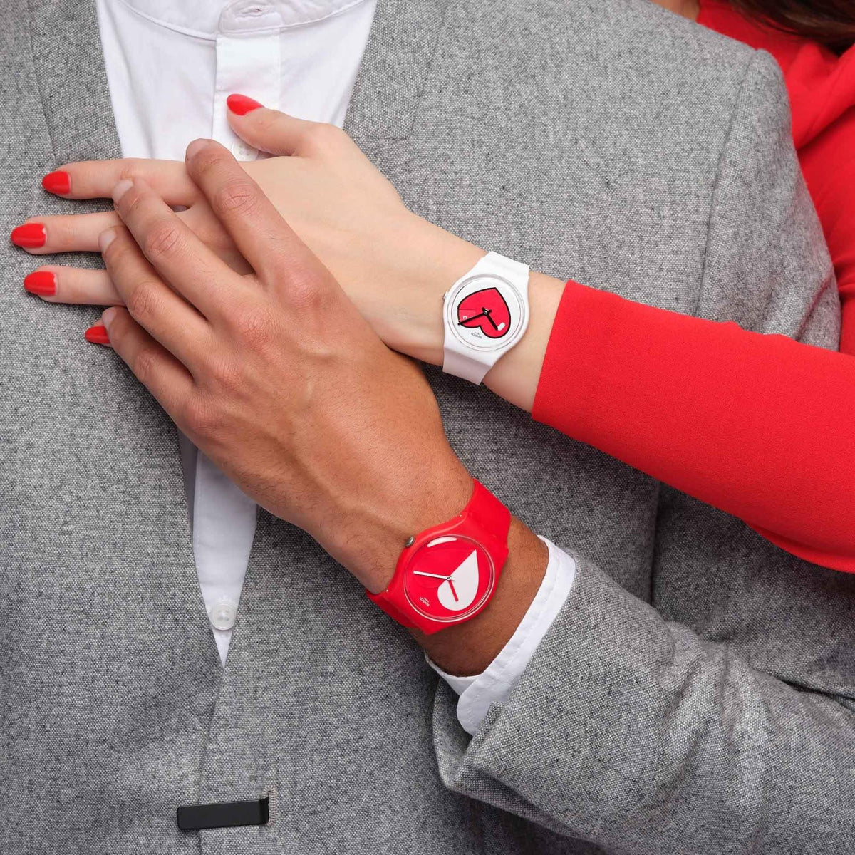 Close-up of a couple's hands, one wears the white Swatch heart watch against a red sleeve, and the other wears a matching red Swatch watch against a grey blazer