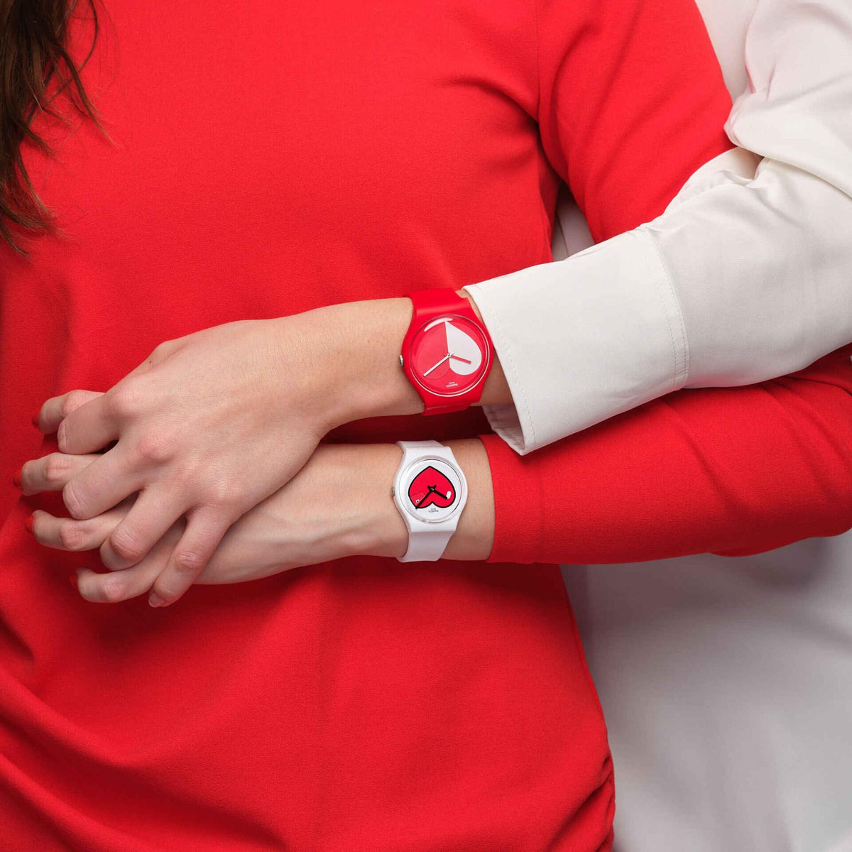 Close-up of a couple with interlocked hands, showcasing the white Swatch Originals Love O'Clock watch on the lower wrist and its matching red counterpart on the upper wrist