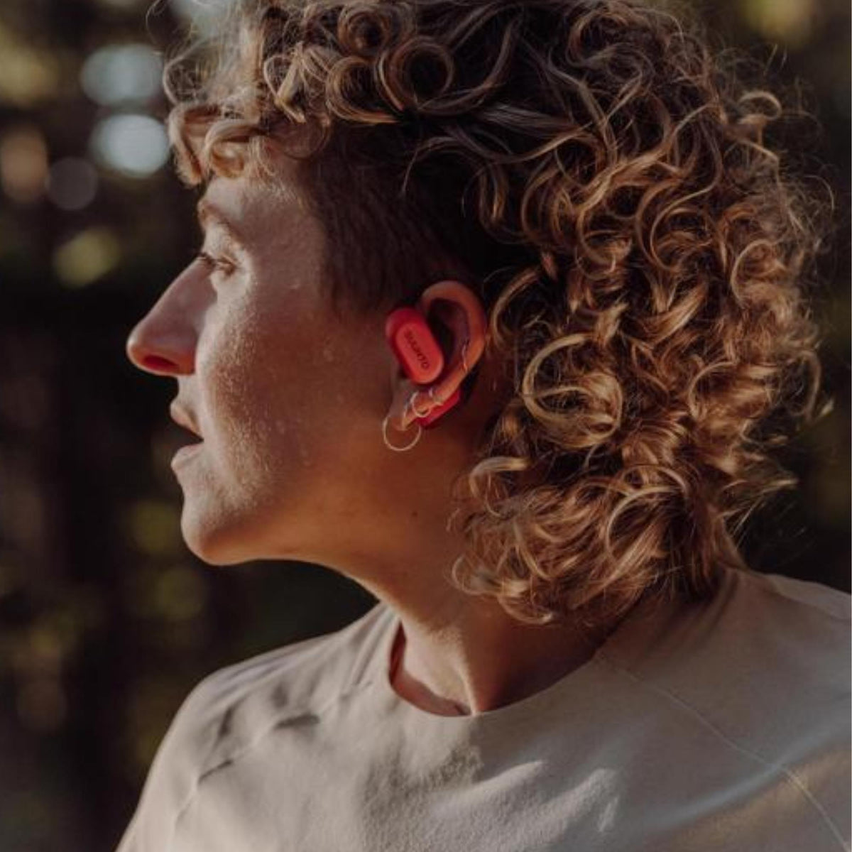 Suunto Spark Open-Ear SS051357000 lifestyle side perspective on female runner wearing coral orange bluetooth earbuds outdoors