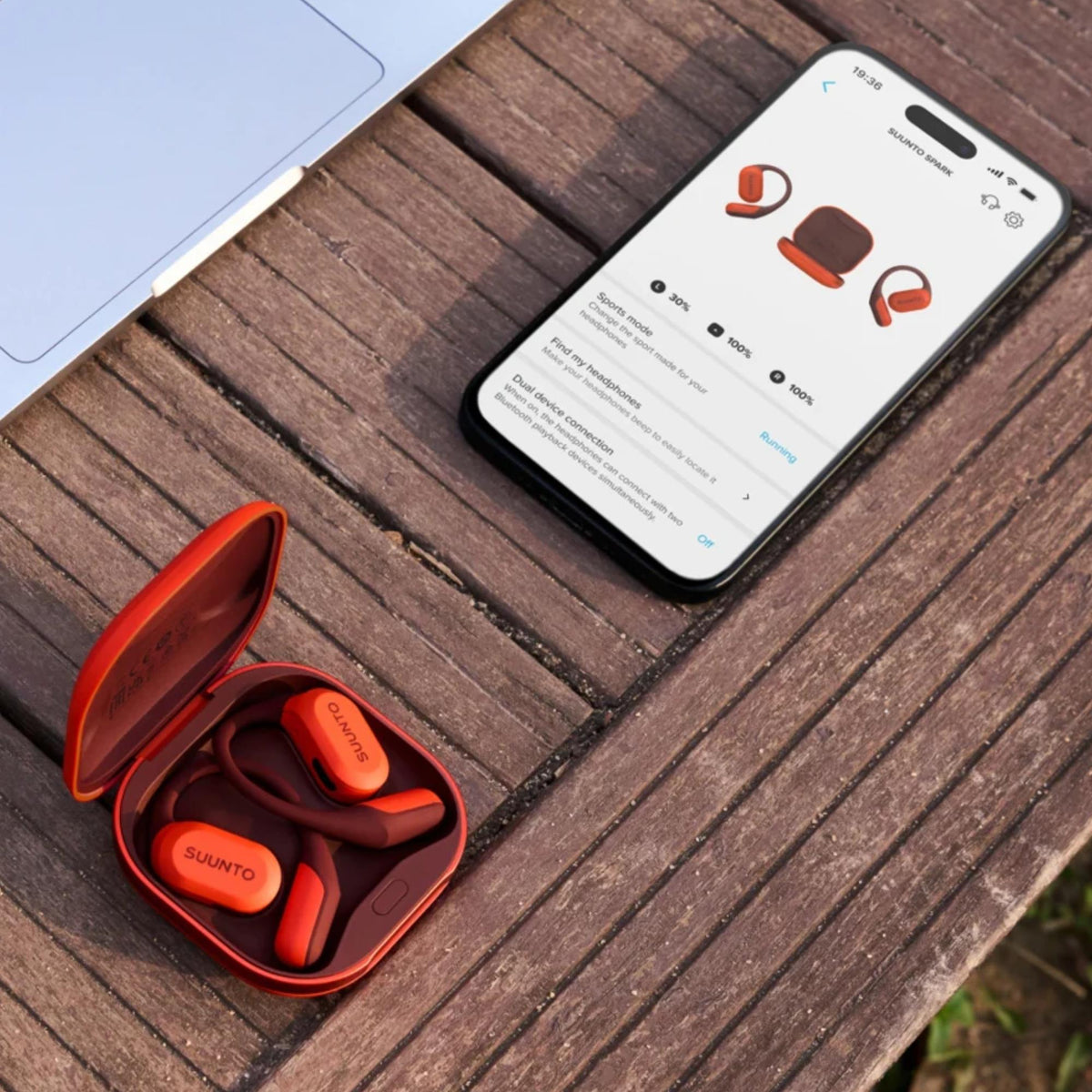 Suunto Spark Open-Ear SS051357000 lifestyle view of open charging case beside smartphone app on wooden table