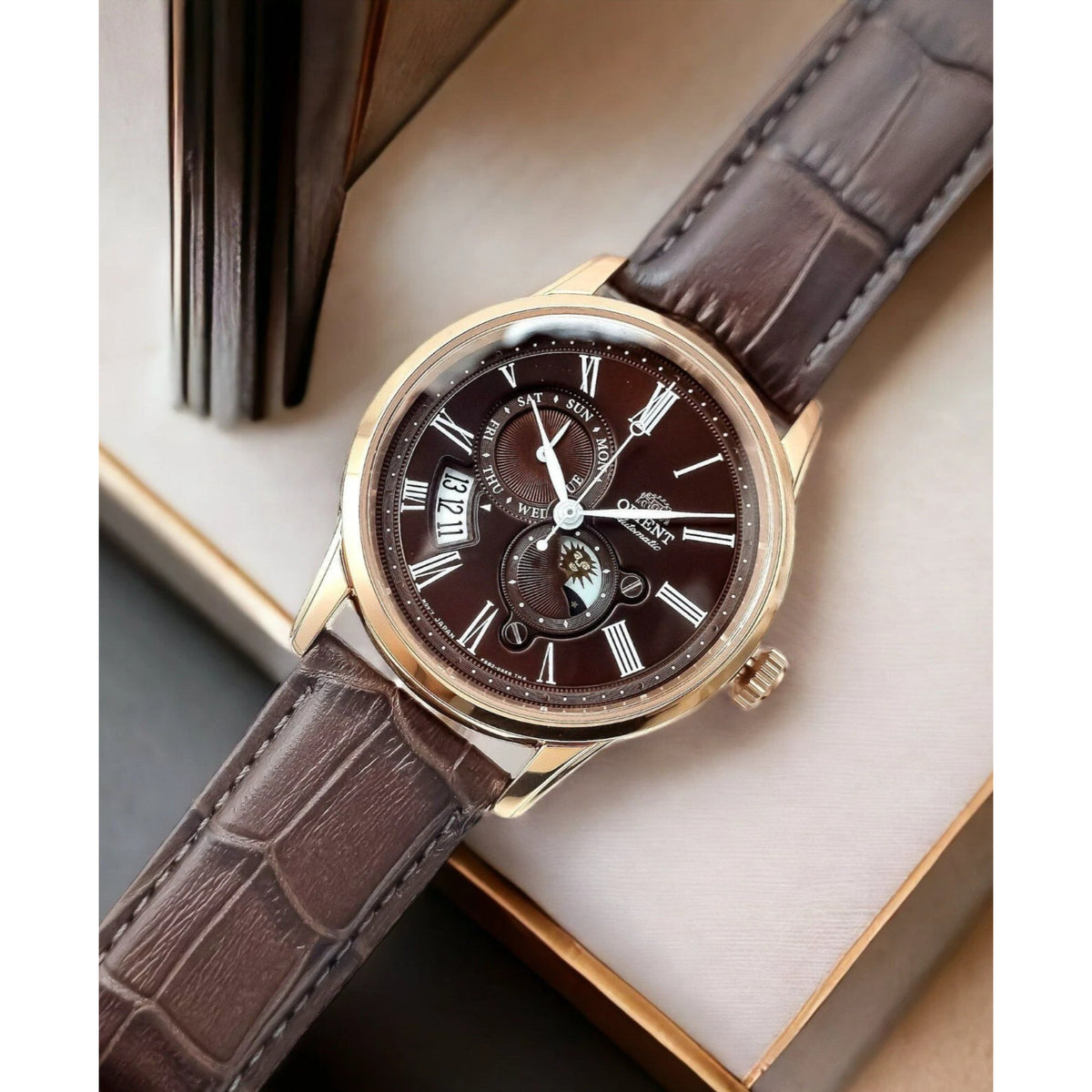 Orient Bambino Sun and Moon RA-AK0009T30B wristwatch on surface, brown leather strap and rose-gold case with dark dial